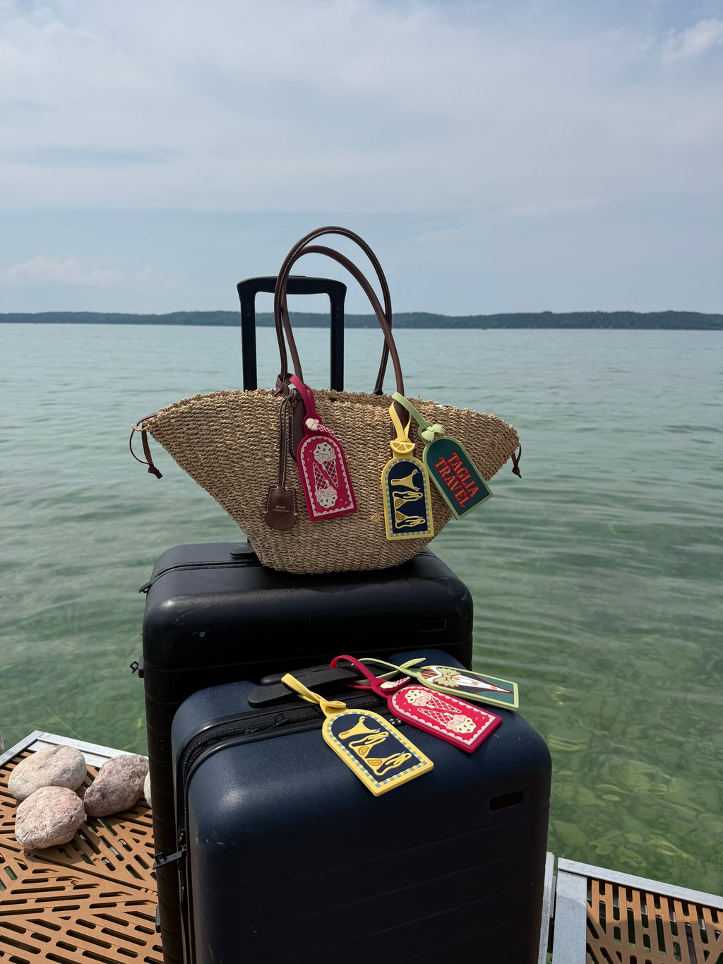 A woven straw tote bag with brown handles and colorful embroidered luggage tags rests on top of stacked black suitcases on a dock, with calm water and distant land under a cloudy sky in the background.