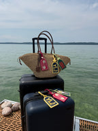 A woven straw tote bag with brown handles and colorful embroidered luggage tags rests on top of stacked black suitcases on a dock, with calm water and distant land under a cloudy sky in the background.