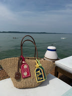 A woven straw tote bag with brown leather handles sits on a white table deck with a lake or sea and distant land visible in the background. Two decorative luggage tags are attached to the bag.