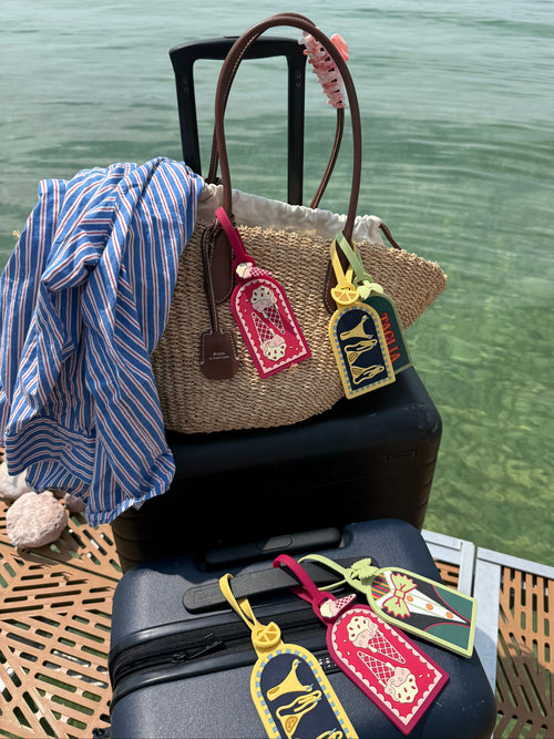 A straw tote bag with brown leather handles and colorful embroidered luggage tags sits on top of stacked black suitcases by the water. A blue-and-white striped shirt is draped over the tote, and a wooden dock and green water are visible in the background.